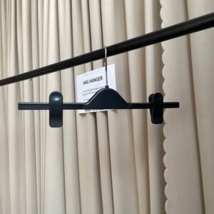 Front view of black plastic pants hangers with adjustable clips, polished chrome swivel hook, hanging on black clothing rail with supplier info card against beige pleated curtain for apparel retail wholesale