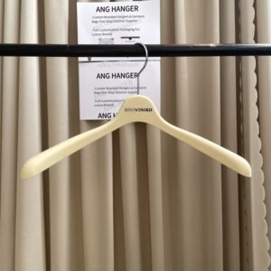 Front view of best shirt hangers in smooth ivory plastic with contoured shoulder design, polished chrome swivel hook and custom printed brand logo, hanging on black clothing rail with two supplier QR cards behind beige pleated curtain for apparel wholesale