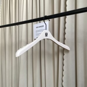 Angled front view of kid hangers with non-slip grip dots and black swivel hook, custom brand emblem on the neck, hanging on black clothing rail with supplier info card against beige pleated curtain for children's apparel wholesale