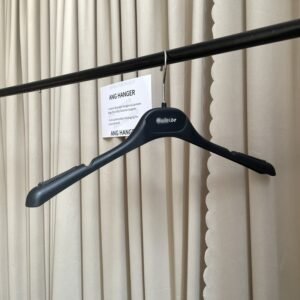 Black non-slip shirt hanger with textured shoulder pads and strap notches, chrome swivel hook with custom brand logo, hanging on clothing rail with supplier card against beige curtain, designed for shirts and wedding dress wholesale