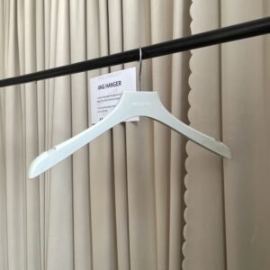 Slim white plastic hanger with integrated strap notches and chrome swivel hook, custom brand logo printed on neck, hanging on clothing rail with supplier info card against beige pleated curtain for apparel wholesale display