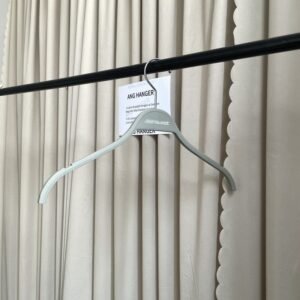 Slanted view of t shirts hangers with built-in strap notches, chrome swivel hook and custom brand marking, hanging on black clothing rail with supplier info card against pleated curtain for apparel retail wholesale