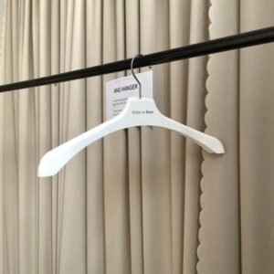 Angled view of smooth white shirt hangers with rounded shoulders, chrome swivel hook and custom brand logo, hanging on black clothing rail with supplier info card against beige pleated curtain for dress shirt retail displa