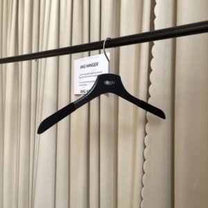 Angled front view of jacket on hanger with velvet non-slip shoulder strips, chrome swivel hook and custom brand logo, hanging on black clothing rail with supplier info card against beige pleated curtain for outerwear retail wholesale