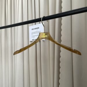 Amber translucent acrylic hanger with polished gold swivel hook and built-in strap notches, custom gold foil brand logo on neck, hanging on black clothing rail with supplier info card against beige pleated curtain for boutique wholesale display