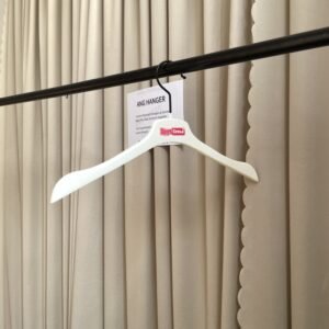 White matte shirt hanger with smooth curved shoulders and black metal swivel hook, custom printed brand logo on neck, hanging on black clothing rail with supplier info card against beige pleated curtain for apparel retail wholesale
