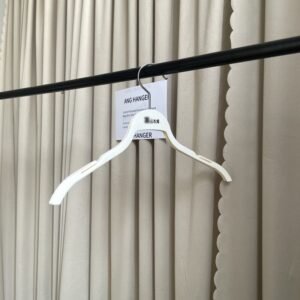 Angled front view of thin hangers with non-slip strap notches, polished chrome swivel hook and custom brand marking, hanging on black clothing rail with supplier info card against beige pleated curtain for apparel retail wholesale