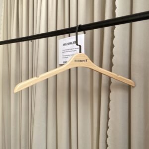 Angled front view of wood-grain plastic clothes hangers with shirt shoulder notches, black swivel hook and custom brand logo, hanging on black clothing rail with supplier info card against beige pleated curtain for apparel retail wholesale