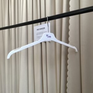 Angled front view of white plastic hangers with non-slip shoulder notches, polished chrome swivel hook and custom brand logo, hanging on black clothing rail with supplier info card against beige pleated curtain for apparel retail wholesale