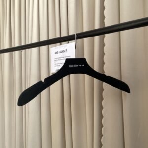 Angled front view of full velvet black plastic hangers with non-slip shoulder grips, polished chrome swivel hook and custom brand logo, hanging on black clothing rail with supplier info card against beige pleated curtain for luxury apparel retail wholesale