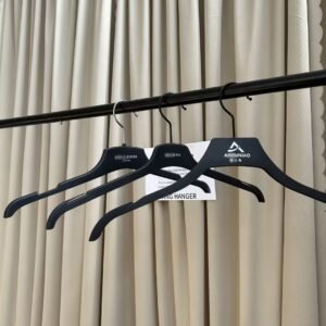 Front view of three matte plastic hangers black with non-slip shoulder notches, mixed chrome and black swivel hooks with custom brand logos, hanging on black clothing rail with supplier info card against beige pleated curtain for apparel wholesale