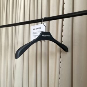 Angled front view of wide shoulder shirt coat hangers with non-slip shoulder texture, polished chrome swivel hook and custom brand logo, hanging on black clothing rail with supplier info card against beige pleated curtain for apparel retail wholesale