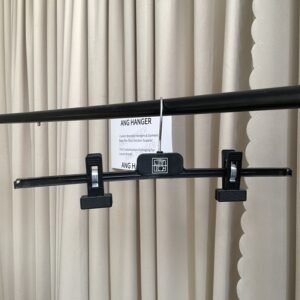 Front view of black plastic hangers with clips, metal nonslip adjustable grips and polished chrome swivel hook, hanging on black clothing rail with supplier info card against beige pleated curtain for apparel retail and wholesale