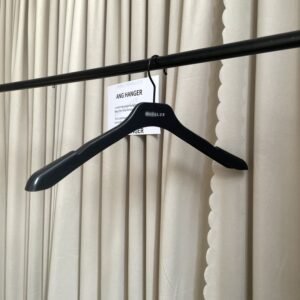 Angled front view of smooth black plastic clothes hangers bulk style with wide shoulders, black swivel hook and custom brand logo, hanging on black clothing rail with supplier info card against beige pleated curtain for apparel wholesale