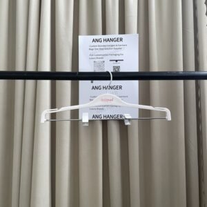 Front view of white 2-in-1 plastic pant hangers with adjustable metal clips, white swivel hook and custom brand logo, hanging on black clothing rail with two QR-code supplier cards against beige pleated curtain for apparel retail and wholesale