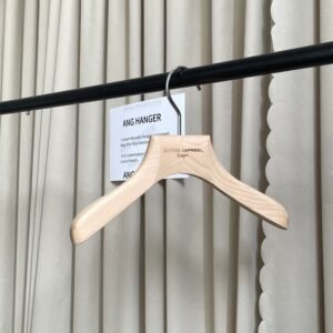 Angled front view of natural beech wooden hangers for shirts with wide shoulder design, polished chrome swivel hook and custom laser engraved logo, hanging on black clothing rail with supplier info card against beige pleated curtain for premium apparel retail