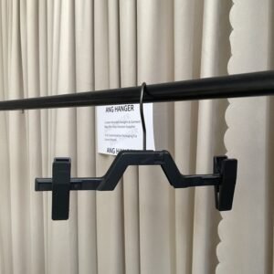 Front view of one-piece black plastic hanger for pants with non-slip clips, polished chrome swivel hook, hanging on black clothing rail with supplier info card against beige pleated curtain for apparel retail and wholesale