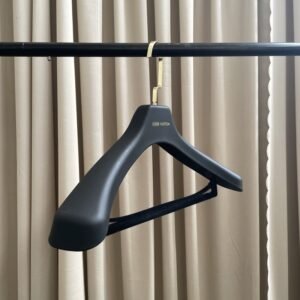 Matte black heavy-duty coat hanger with gold flat hook and velvet non-slip trouser bar, custom printed brand logo, hanging on clothing rack with supplier cards for luxury apparel wholesale