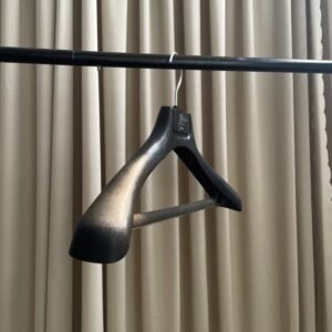 Textured plastic black coat hanger with chrome swivel hook and anti-slip trouser bar, custom logo plate on top, hanging on clothing rod against pleated beige curtain for apparel wholesale display